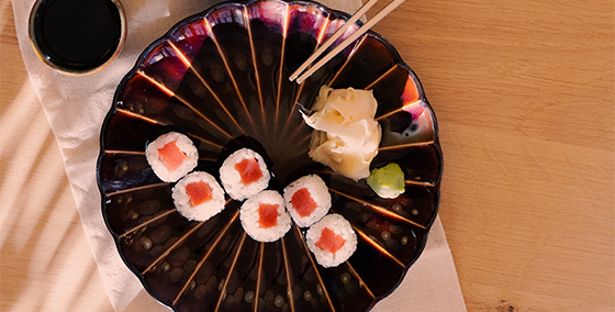Plant-based sushi alternatives Raw TunaliciOus and Raw SalmOnderful from OLALA! beautifully presented on a dark, shell-shaped ceramic plate, offering a vegan twist to traditional sushi. The dish is complemented with ginger, wasabi, and soy sauce, capturing the essence of sustainable dining on a warm wooden backdrop.