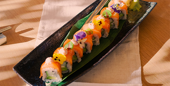 Plant-based sushi rolls, the Raw SalmOnderful by OLALA!, artistically garnished with edible flowers, served on a sleek black plate over a bamboo leaf. The vegan alternative boasts vibrant colors and a fresh appeal, ideal for diners seeking delicious oh lala veggie sushi experiences, presented in an inviting atmosphere with lit candles and soft natural lighting